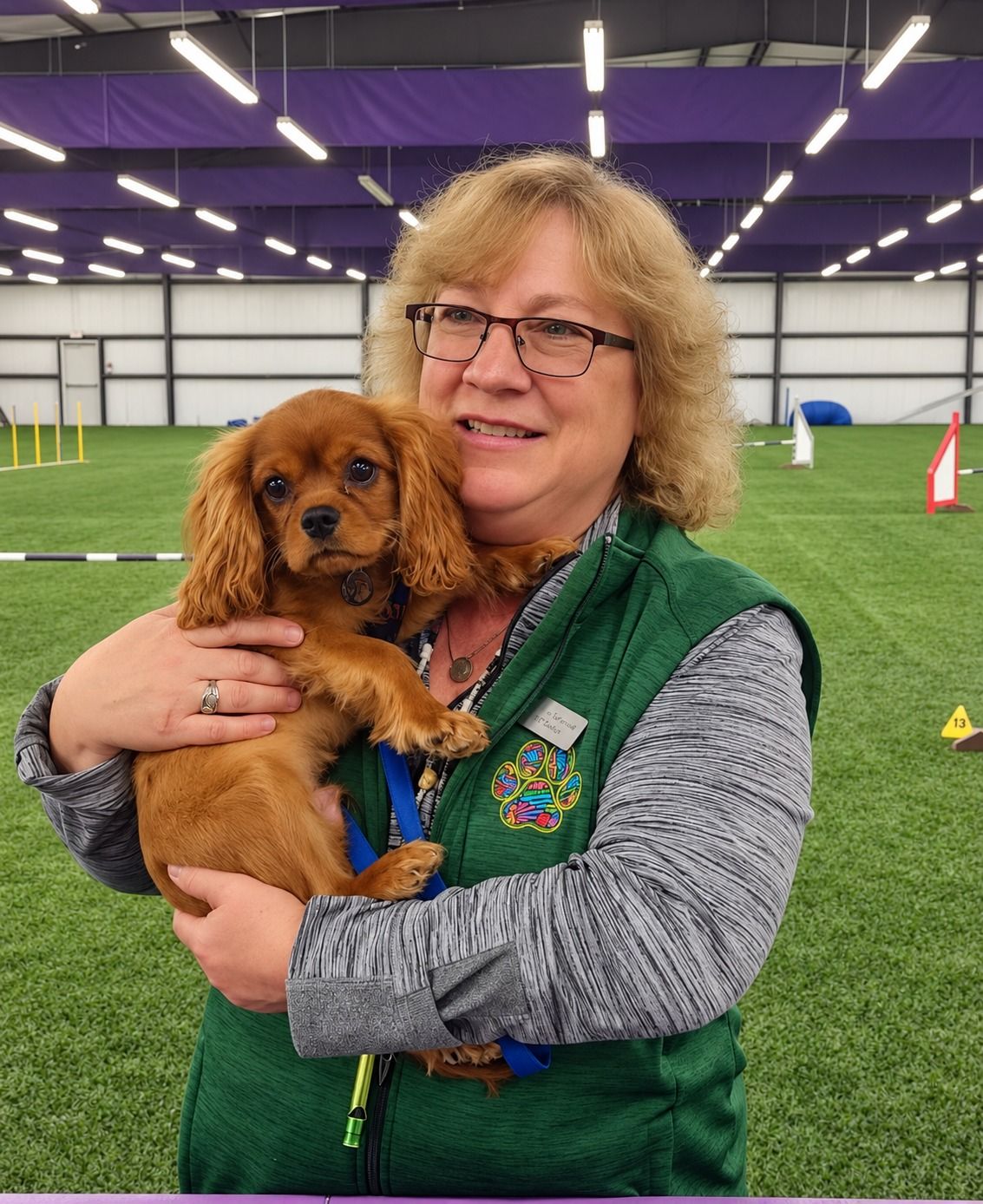 Kristine Schmidt holding a Cavalier King Charles Spaniel in an agility facility.